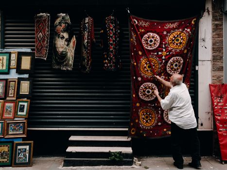 A man hangs vibrant red tapestries beside framed art on an Istanbul street.