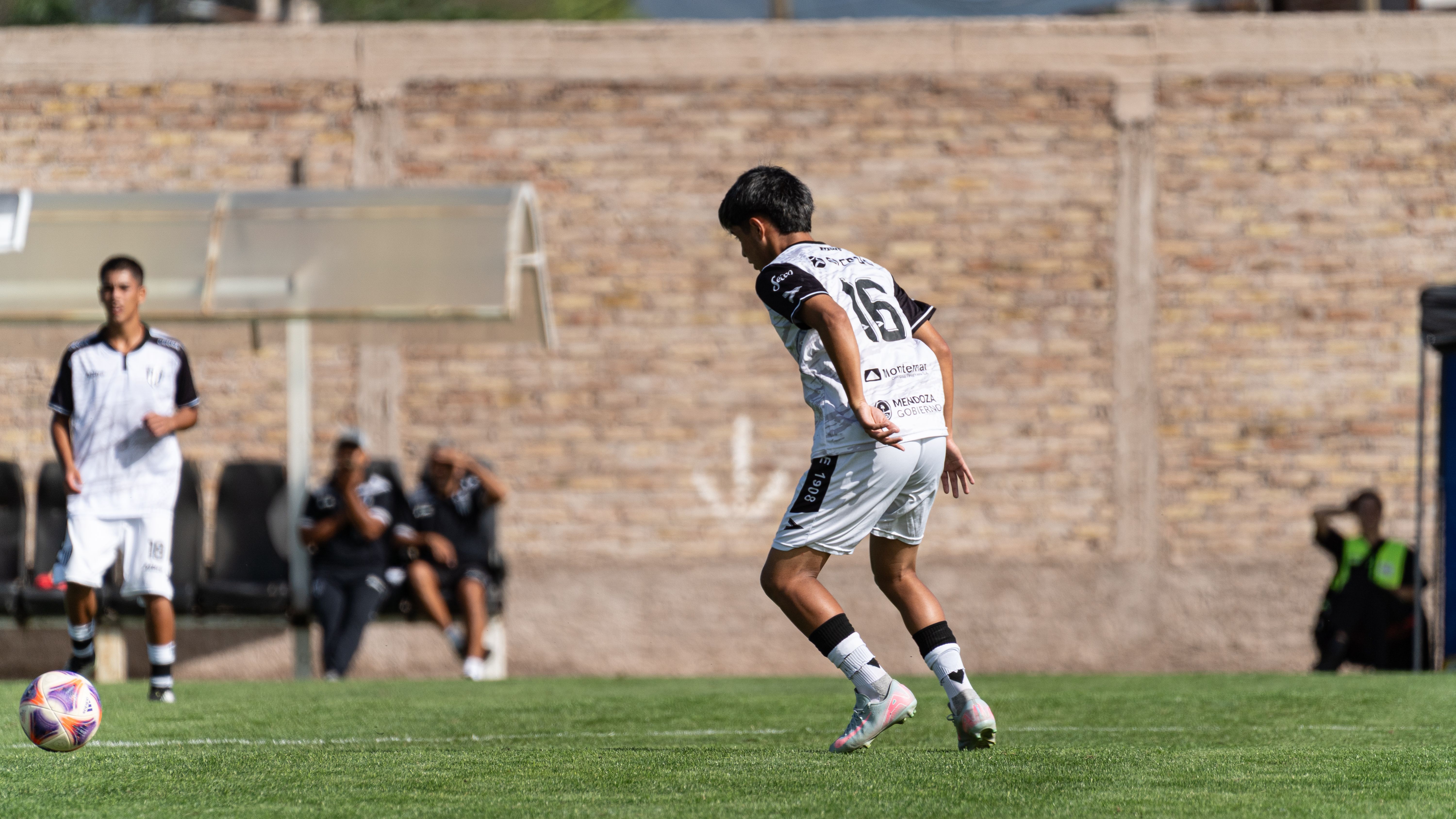 Youth soccer player in action during an outdoor match on a sunny day with a brick wall backdrop.