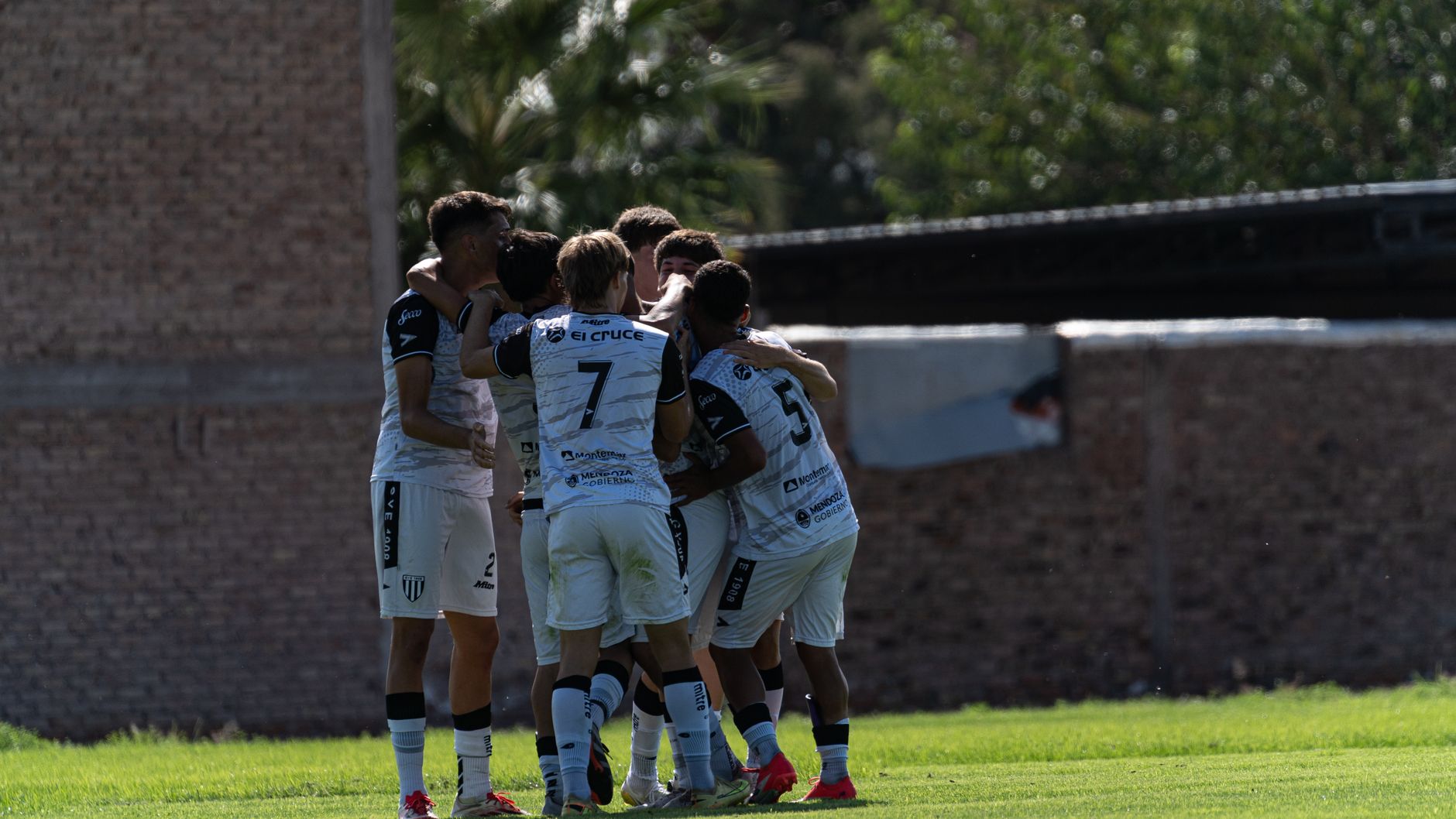 A group of young soccer players huddles in celebration on a sunny day outdoors.