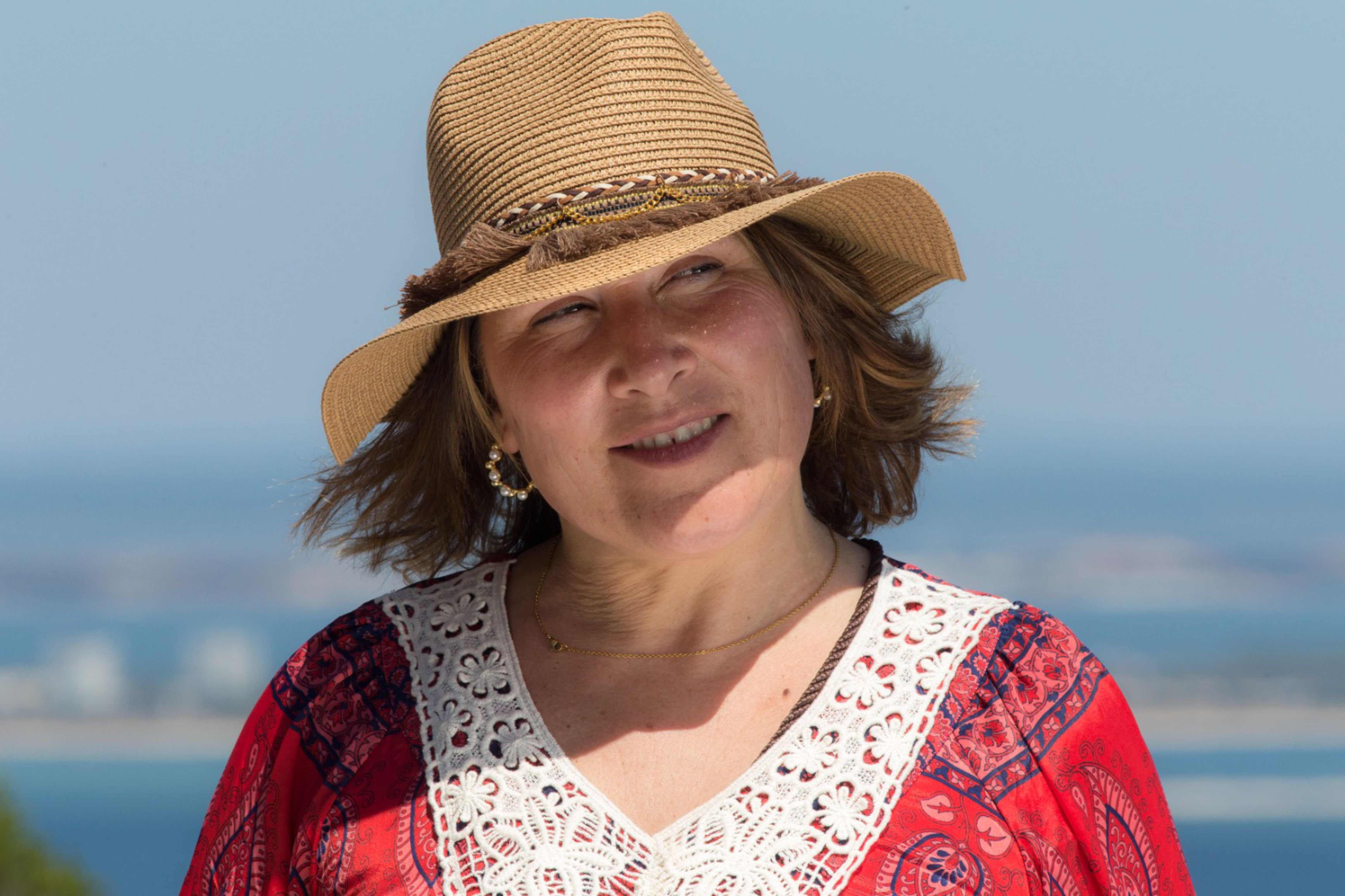 Woman in a straw sun hat and red top smiling outdoors against a clear sky.