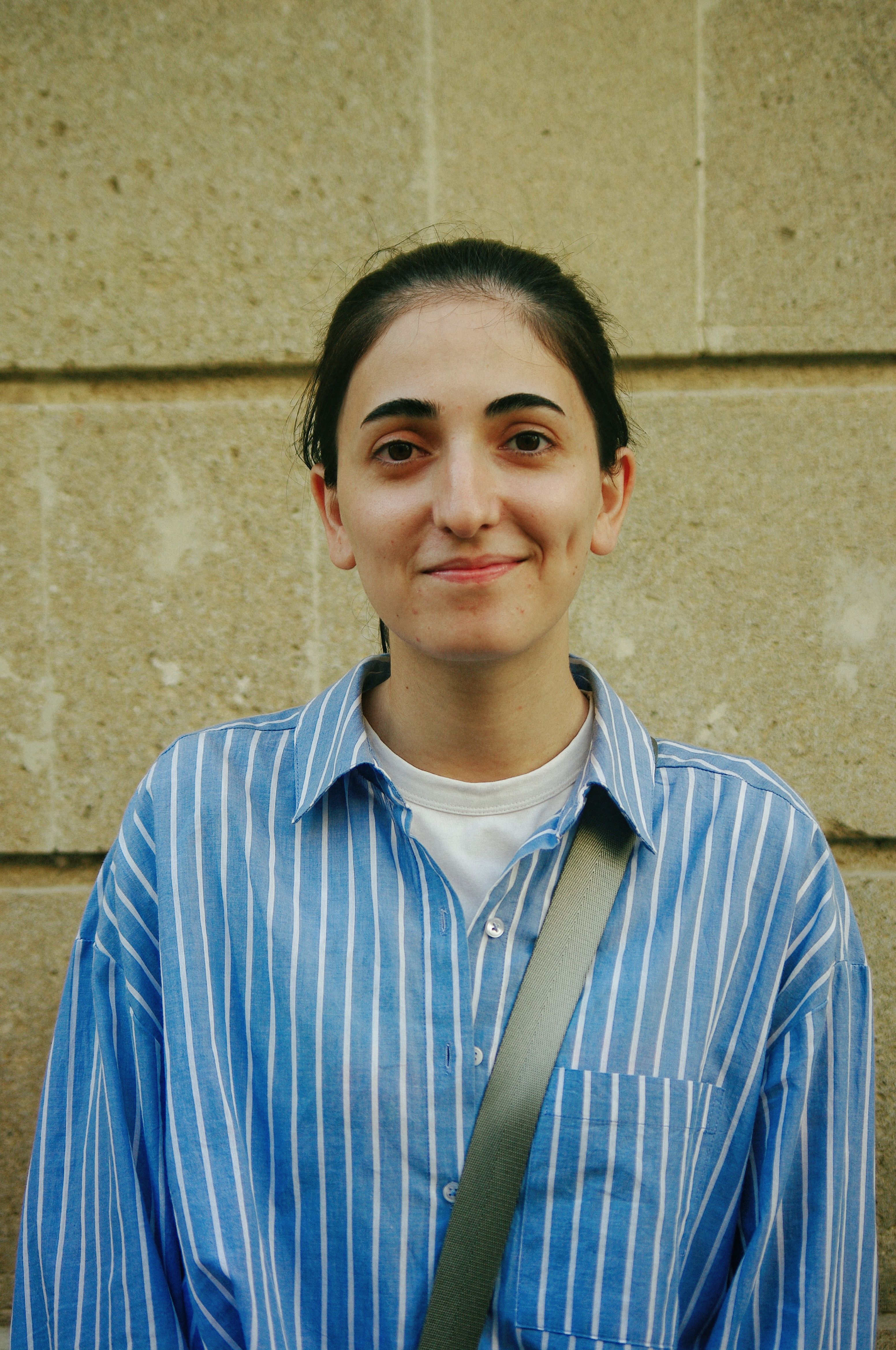 A young adult smiling in a blue striped shirt against a stone wall background.