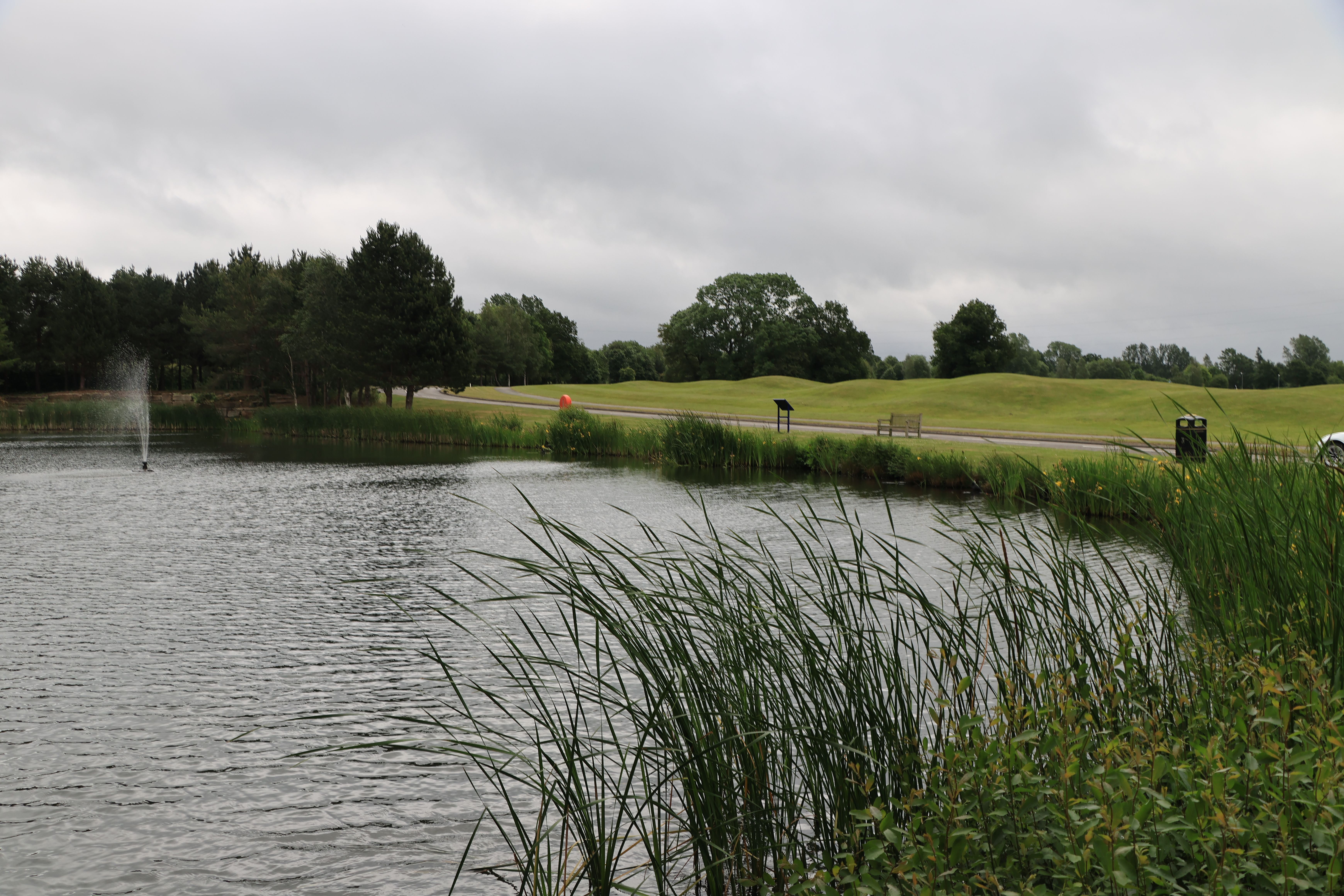 Tranquil lake in Weston, England with fountain and lush greenery under a cloudy sky.