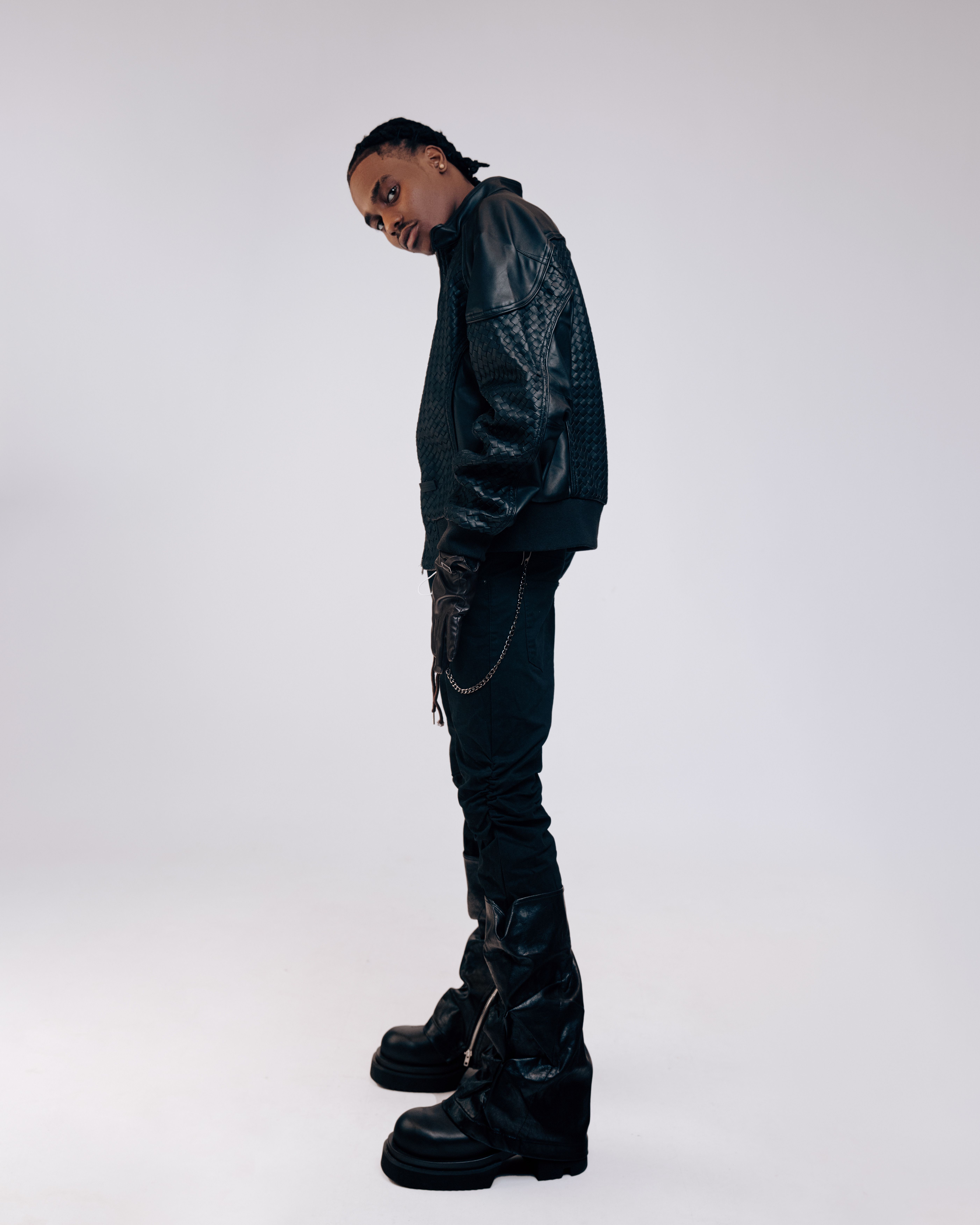 Stylish individual posing in black leather jacket and boots against a white background.