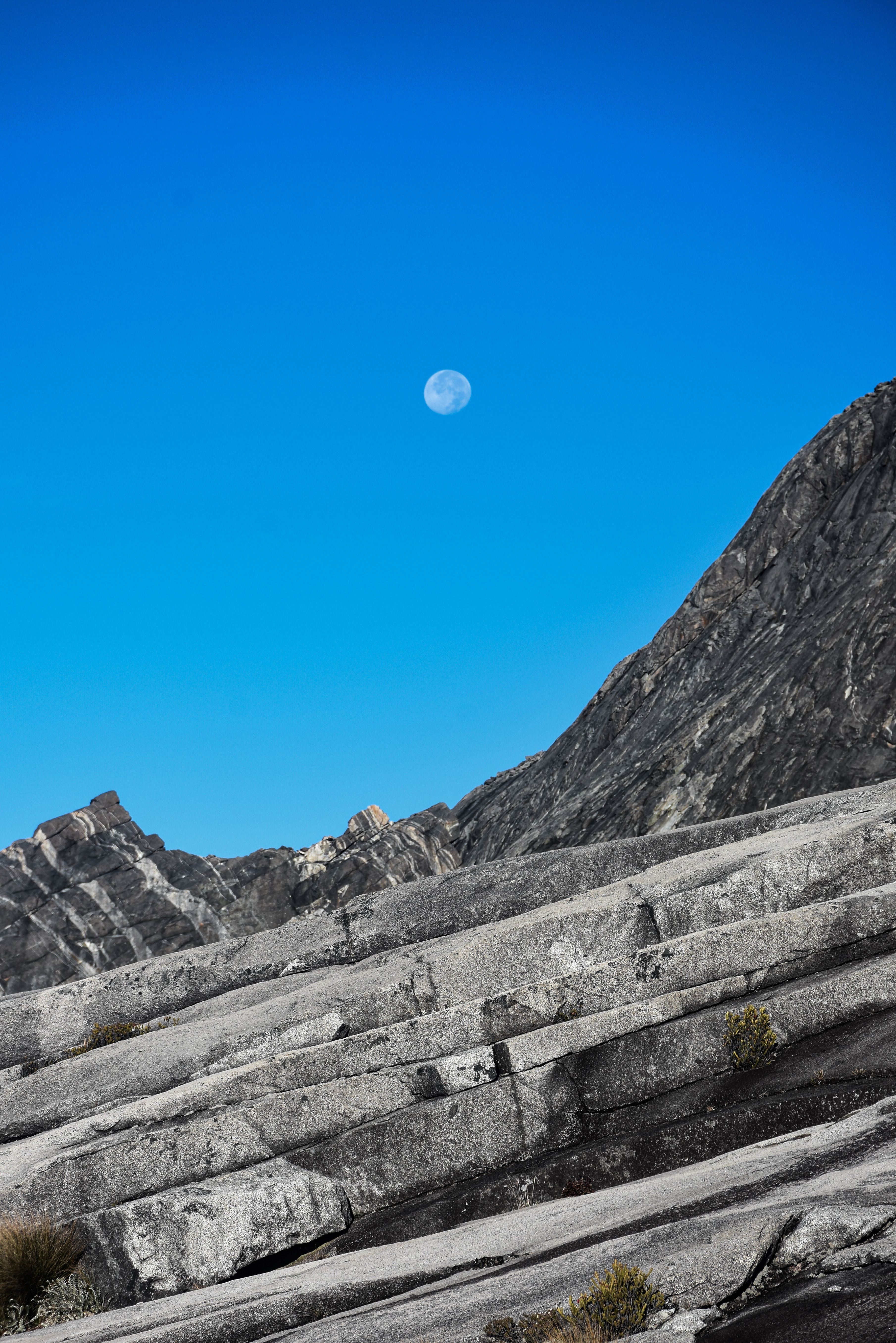 Stunning view of Mount Kinabalu's rocky summit with a clear blue sky and visible moon in Sabah, Malaysia.