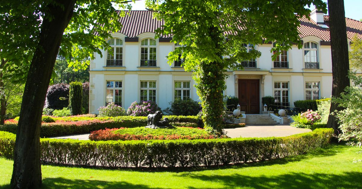 Stately white mansion with manicured gardens and lush trees under a bright sky.