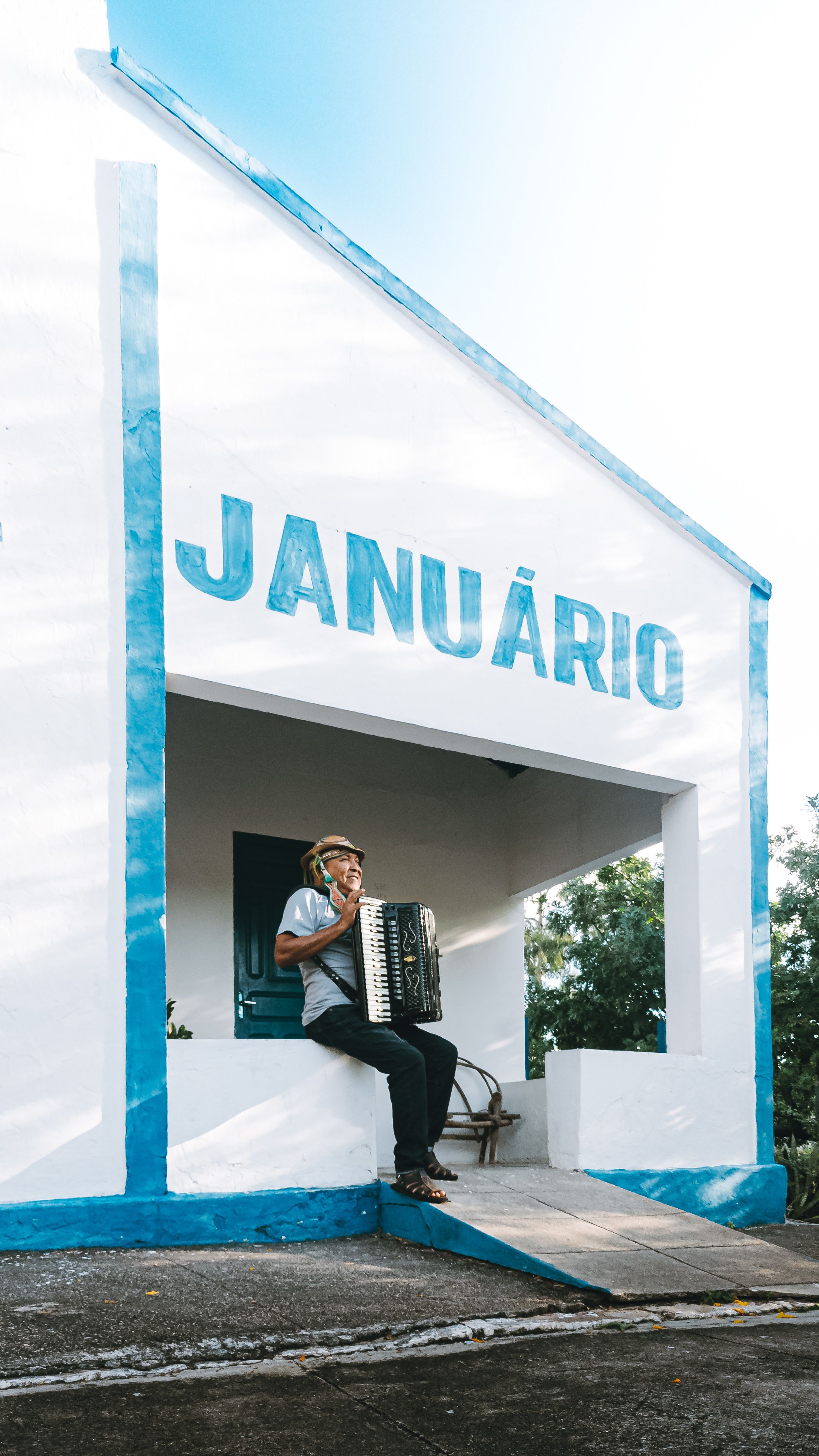 A man sits outside a white building labeled 'Januário', playing an accordion.