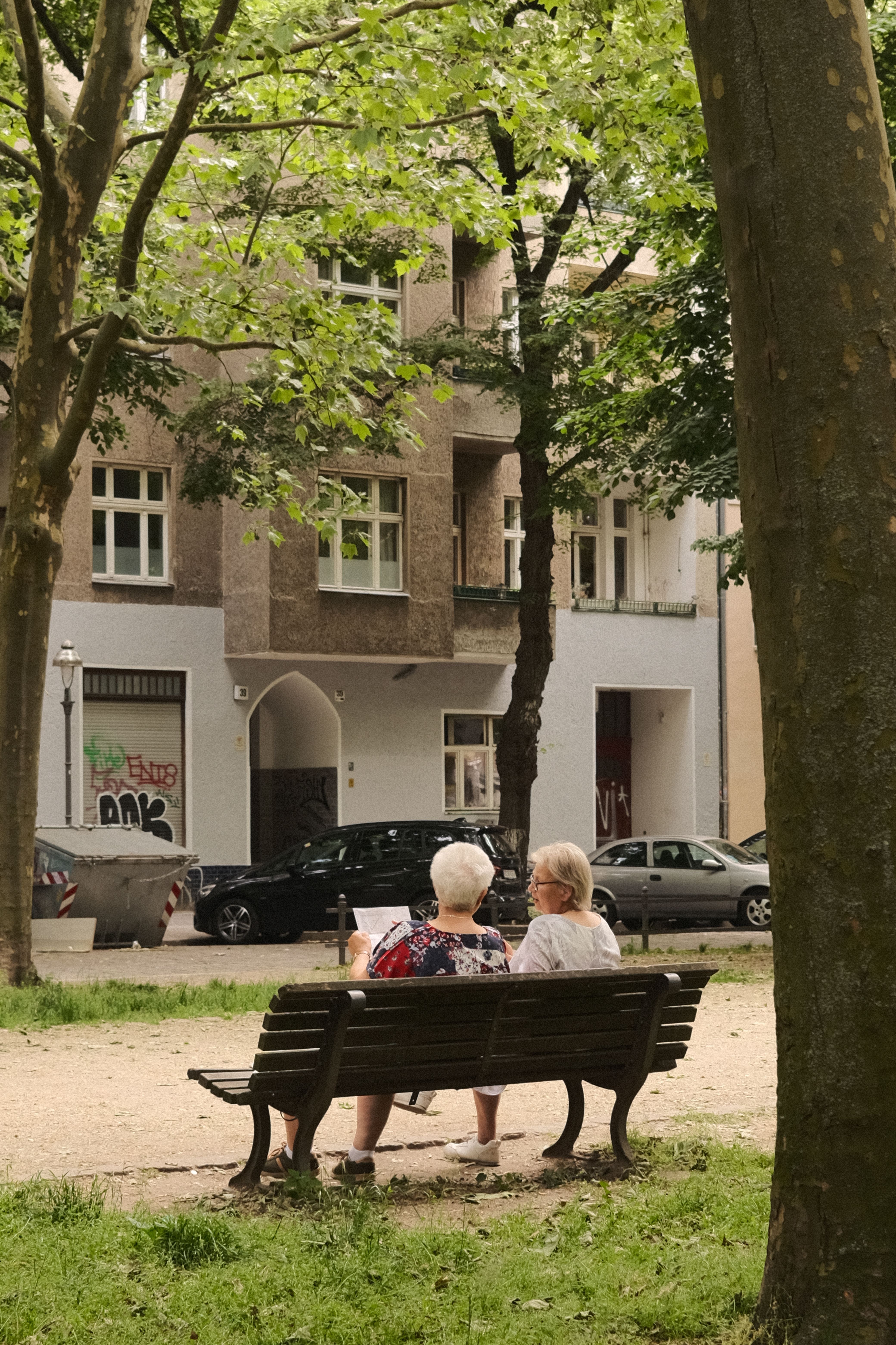 Two senior women seated on a bench in a Berlin park, embracing a tranquil summer day.