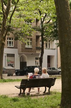 Two senior women seated on a bench in a Berlin park, embracing a tranquil summer day.