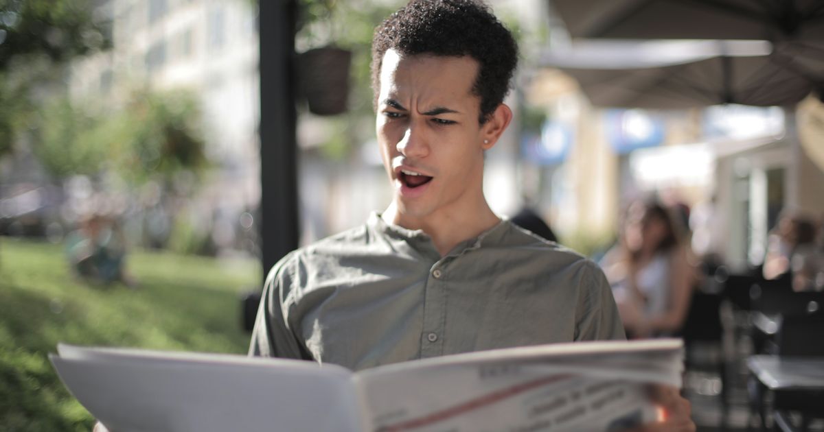 A young man looks surprised while reading a newspaper at an outdoor cafe
