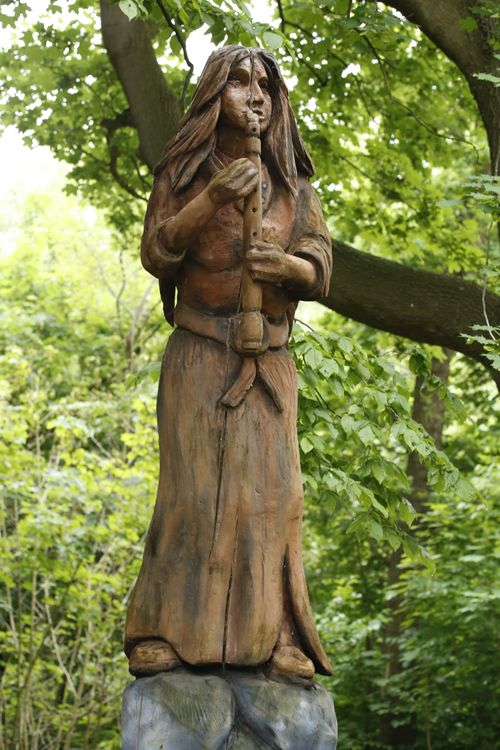 Free Wooden sculpture of a woman playing a flute in a lush forest environment. Stock Photo