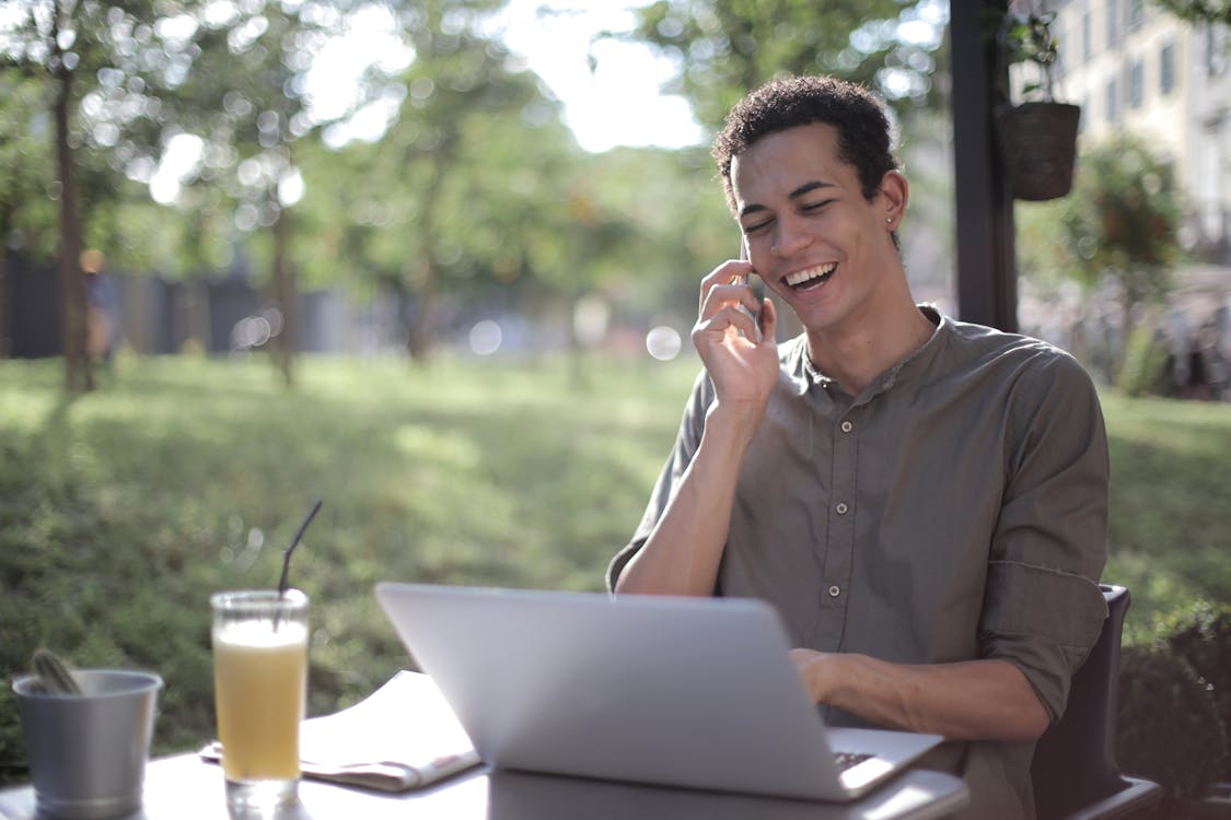 Free Content African American male freelancer wearing casual clothes sitting at table in street cafe with laptop and speaking on cellphone while working on startup Stock Photo