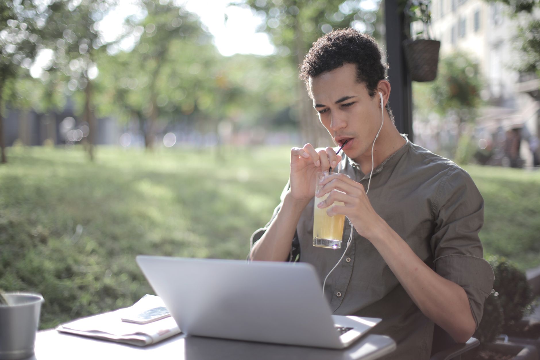 Young African American male in casual wear sipping lemonade through straw and watching video on netbook while sitting in earphones at table in street cafe and chilling during weekend