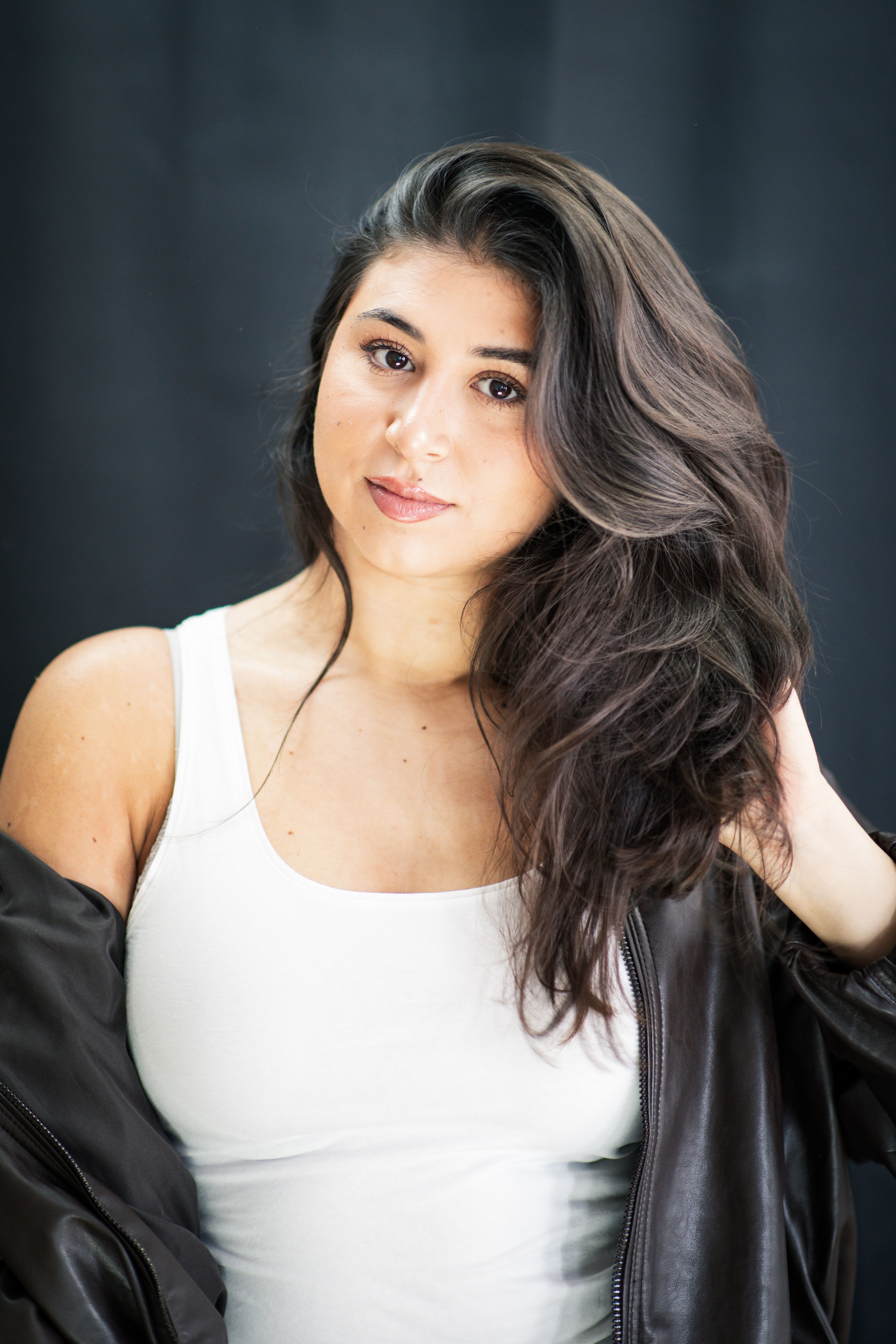 Stylish portrait of a woman wearing a white tank top and leather jacket against a dark background.