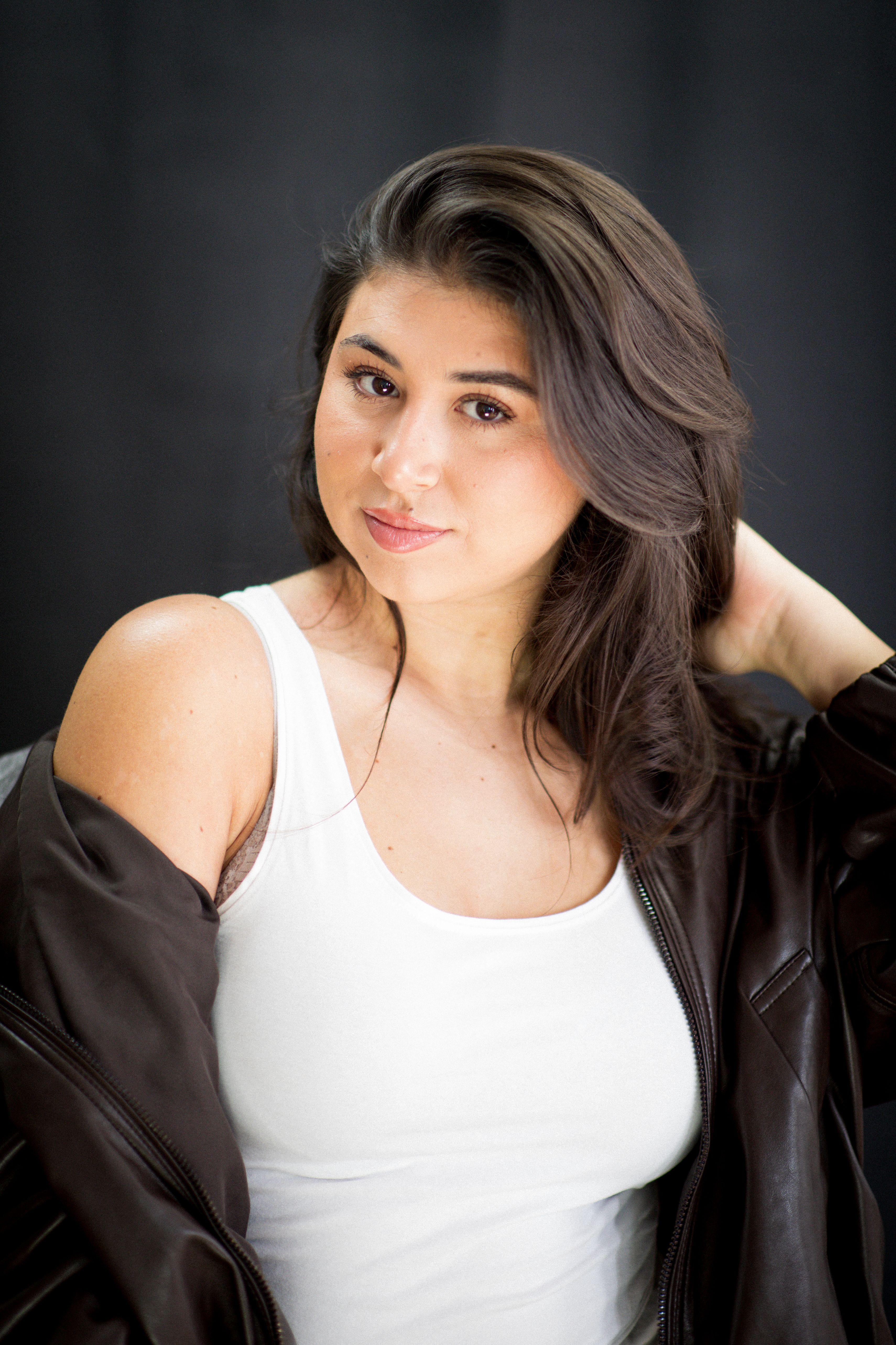 Stylish portrait of a woman wearing a leather jacket over a white tank top against a dark background.