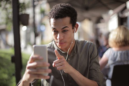 Free Smiling African American male in casual clothing sitting in street cafe and talking via video chat while chilling in city and using mobile phone Stock Photo