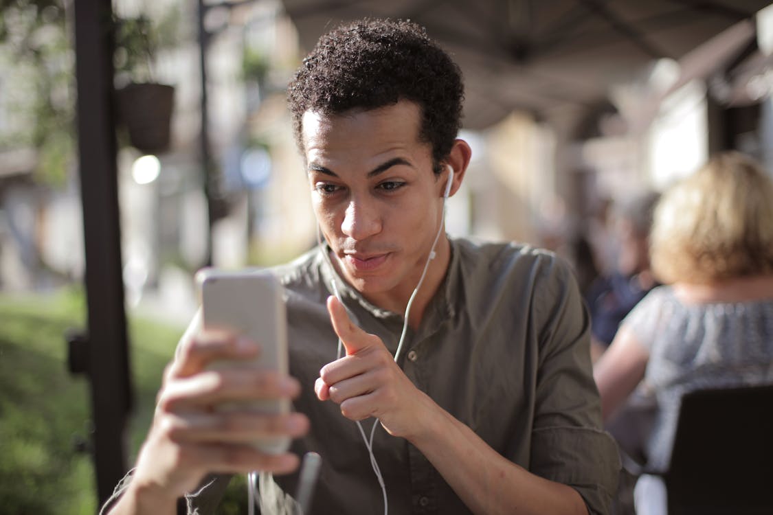 Smiling African American male in casual clothing sitting in street cafe and talking via video chat while chilling in city and using mobile phone