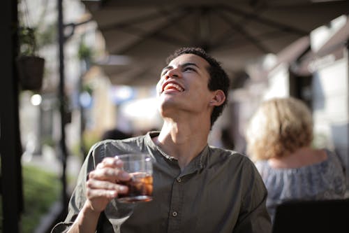 From below joyful young African American male with open mouth in casual clothes looking up while having refreshment with cool tasty beverage on cafe terrace