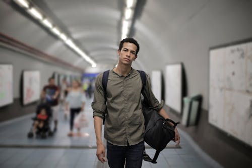 Calm young African American male in casual clothes with big black bag and backpack looking at camera while walking along corridor of underground station against blurred passengers
