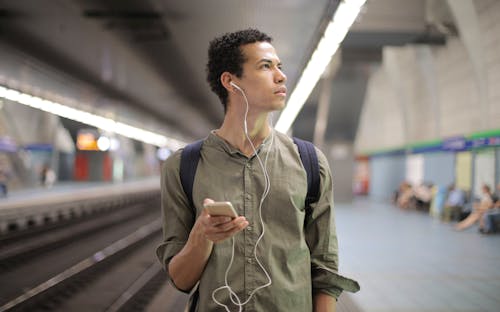 Молодой этнический мужчина в наушниках слушает музыку в ожидании транспорта на современной станции метро