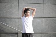 Calm pensive African American sportsman in earphones and sportswear looking away and thinking while stretching arms and listening to music with pleasure on street against gray exterior of contemporary buildings
