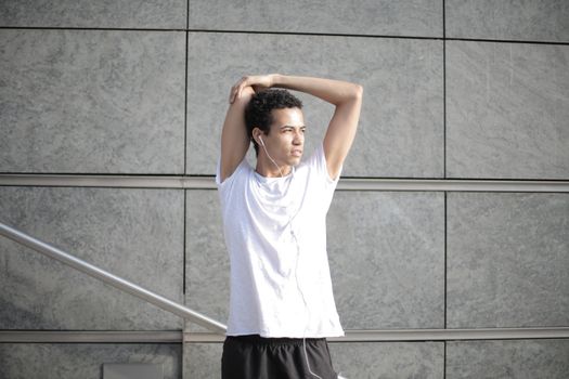 Calm pensive African American sportsman in earphones and sportswear looking away and thinking while stretching arms and listening to music with pleasure on street against gray exterior of contemporary buildings