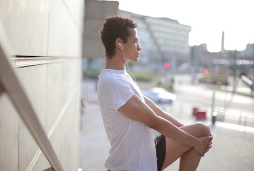 Free Side view of calm thoughtful African American male athlete in earphones and sportswear looking away and stretching leg while standing alone and listening to music on street near modern building in city Stock Photo
