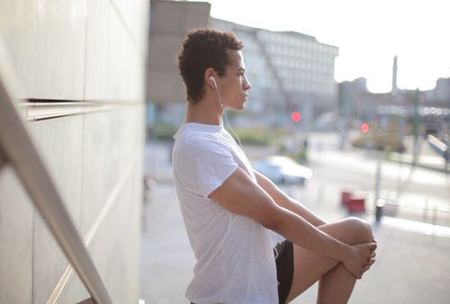 Free Side view of calm thoughtful African American male athlete in earphones and sportswear looking away and stretching leg while standing alone and listening to music on street near modern building in city Stock Photo