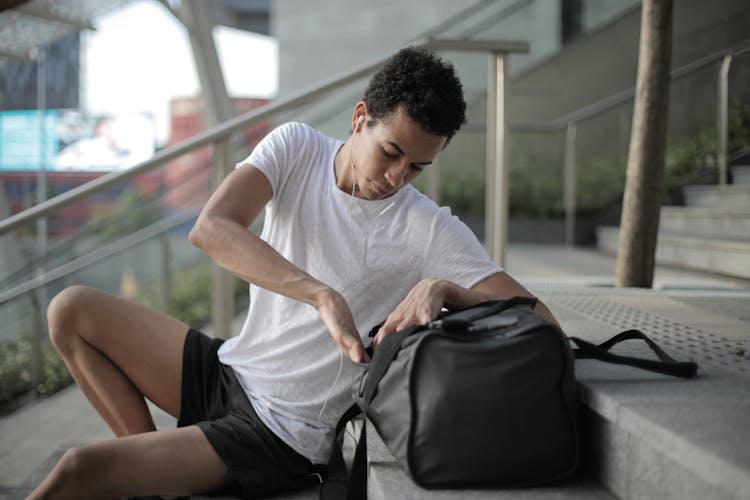 Pensive Millennial Ethnic Male Athlete With Big Sports Bag Listening To Music On Stairs In City