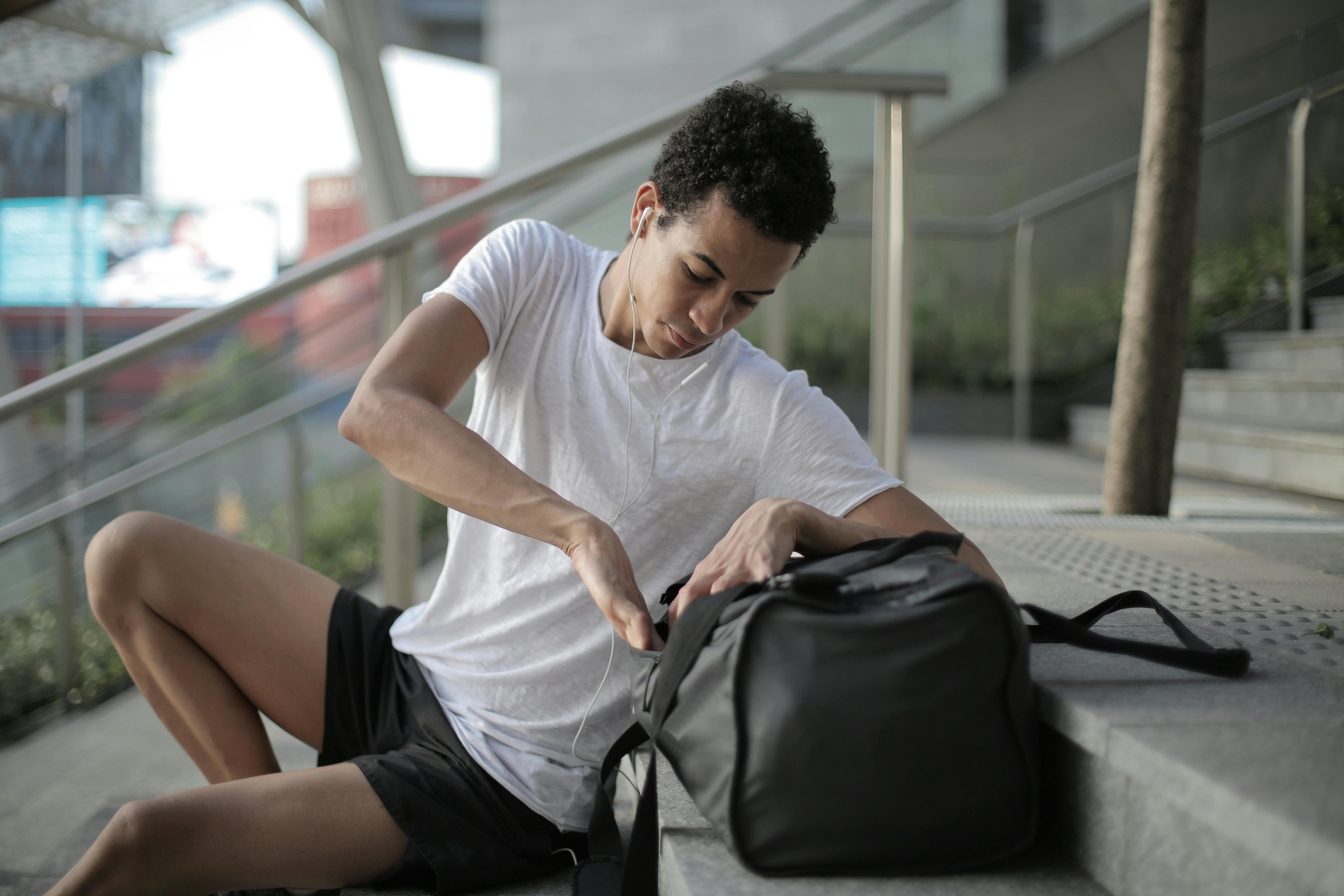 Pensive millennial ethnic male athlete with big sports bag listening to ...