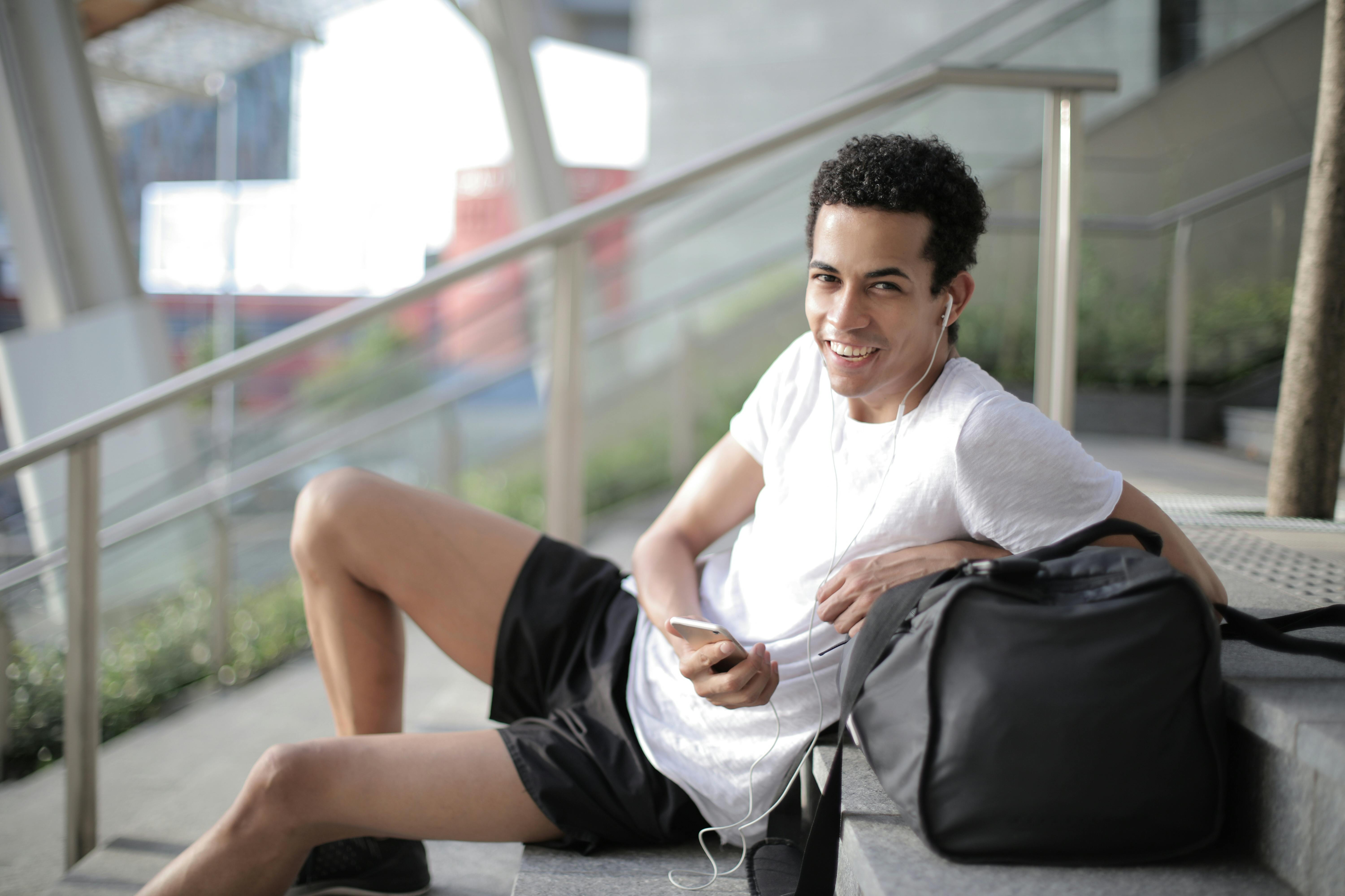 cheerful young ethnic sportsman in earbuds listening to music and using smartphone while resting after training on stairs in city