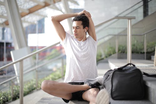 Young ethnic athlete in earbuds stretching body while sitting on stairs at entrance of contemporary building