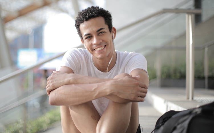 Happy Millennial Ethnic Man In Earphones Listening To Music Lounging On Stairs In City