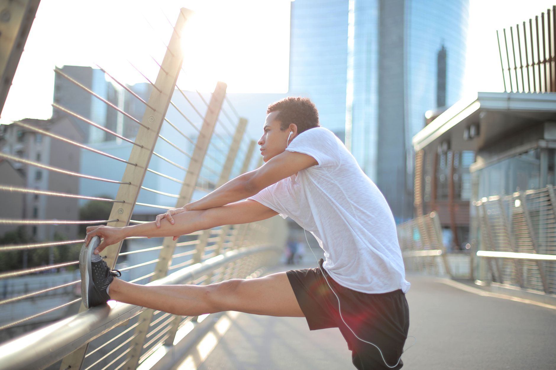 Low angle side view of focused African American male athlete in earphones and sportswear listening to music and stretching body while warming up on street against blurred high rise buildings in downtown