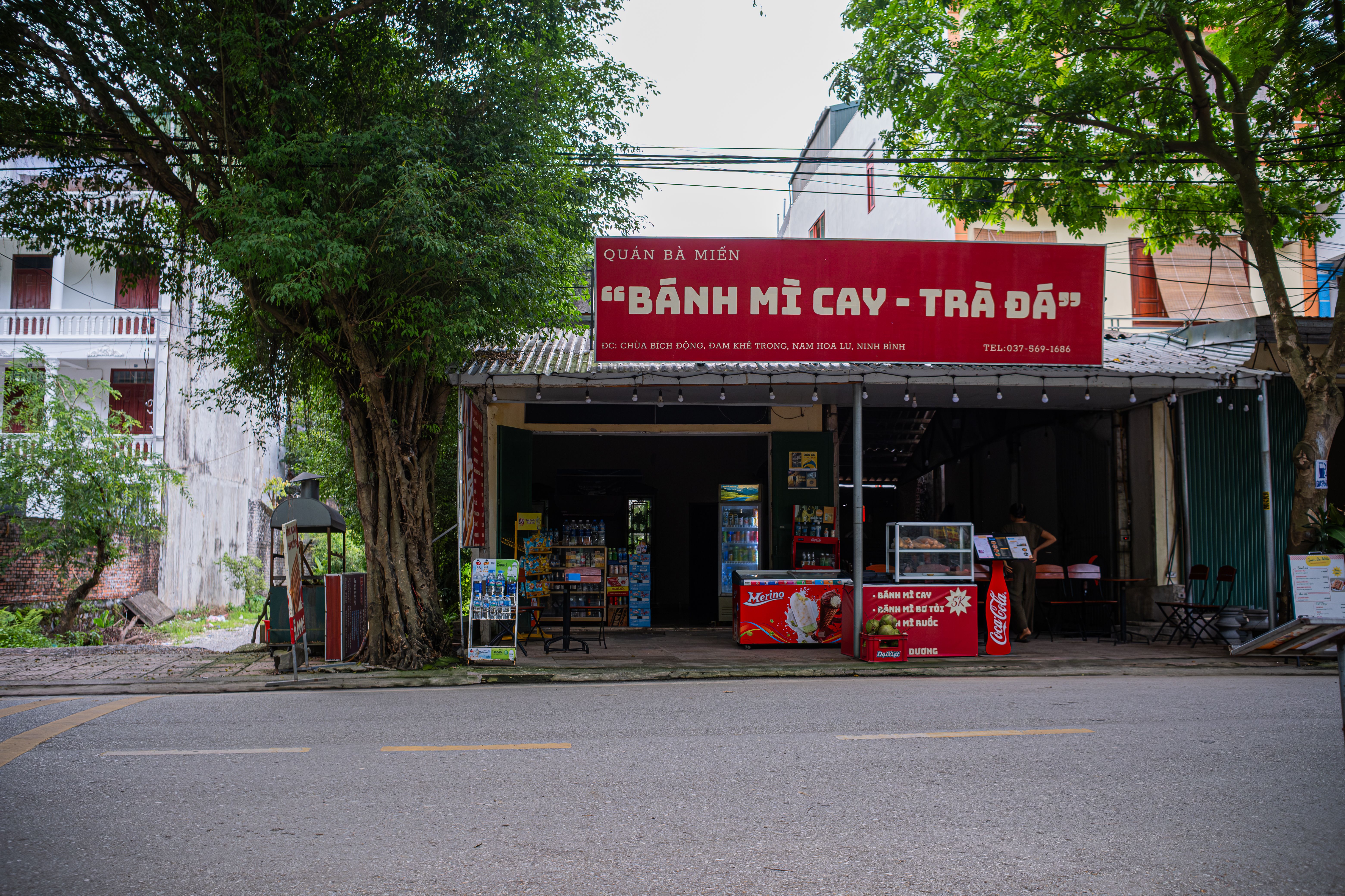 Street vendor shop in Hanoi selling Bánh Mì Cay and Trà Đá under a shady tree.