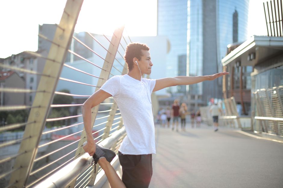 Low angle serious concentrated African American man in earphones wearing white shirt and black shorts stretching legs and listening to music with pleasure on street against blurred urban environment