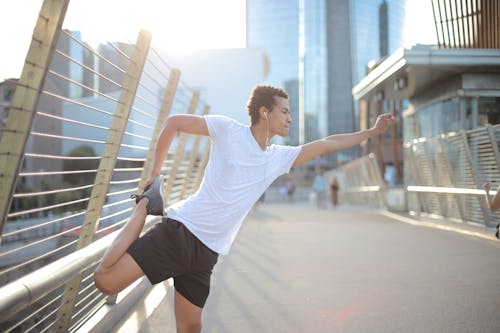 Young African American athlete in earbuds listening to music and stretching legs while training alone on street