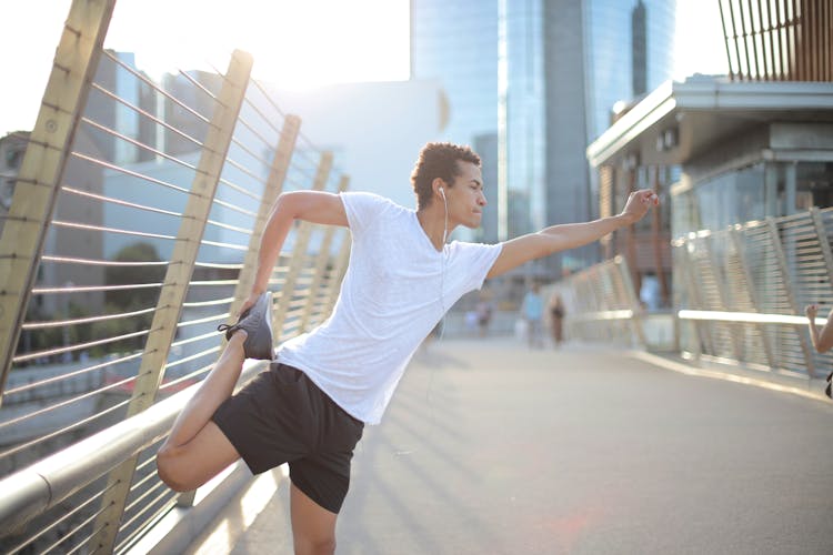 Young African American Athlete In Earbuds Listening To Music And Stretching Legs While Training Alone On Street