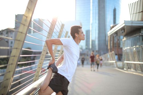 Free Young African American sportsman in earbuds and sportswear listening to music and warming up stretching legs on street against blurred high rise buildings in city Stock Photo
