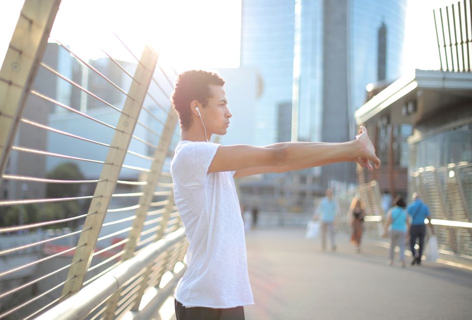 Focused African American man in earbuds and sportswear listening to music and stretching arms while training alone on street against blurred high rise buildings in downtown Focused African American man in earbuds and sportswear listening to music and stretching arms while training alone on street against blurred high rise buildings in downtown