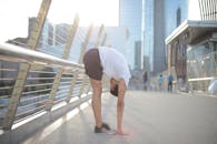 一名戴着耳机、身穿运动服的年轻非裔美国男子侧身站在街上，一边听音乐，一边伸展身体，对着市中心模糊的高层建筑做站立前屈运动