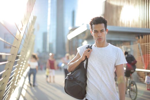 Cool confident African American male in earbuds and casual clothes looking at camera with interest while standing on street with large black bag on shoulder and listening to music against blurred people and urban environment in city