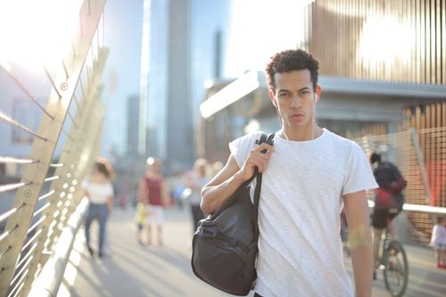 Free Cool confident African American male in earbuds and casual clothes looking at camera with interest while standing on street with large black bag on shoulder and listening to music against blurred people and urban environment in city Stock Photo