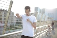 Low angle of positive confident African American male athlete with mouth opened in earphones and sports clothes looking away and doing kick gesture while warming up before training on street against blurred urban environment in sunny day