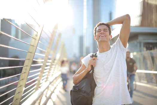 Low angle of overjoyed African American guy in earbuds and casual clothes touching head and laughing while standing with eyes closed on street and listening to music against blurred high rise buildings in downtown