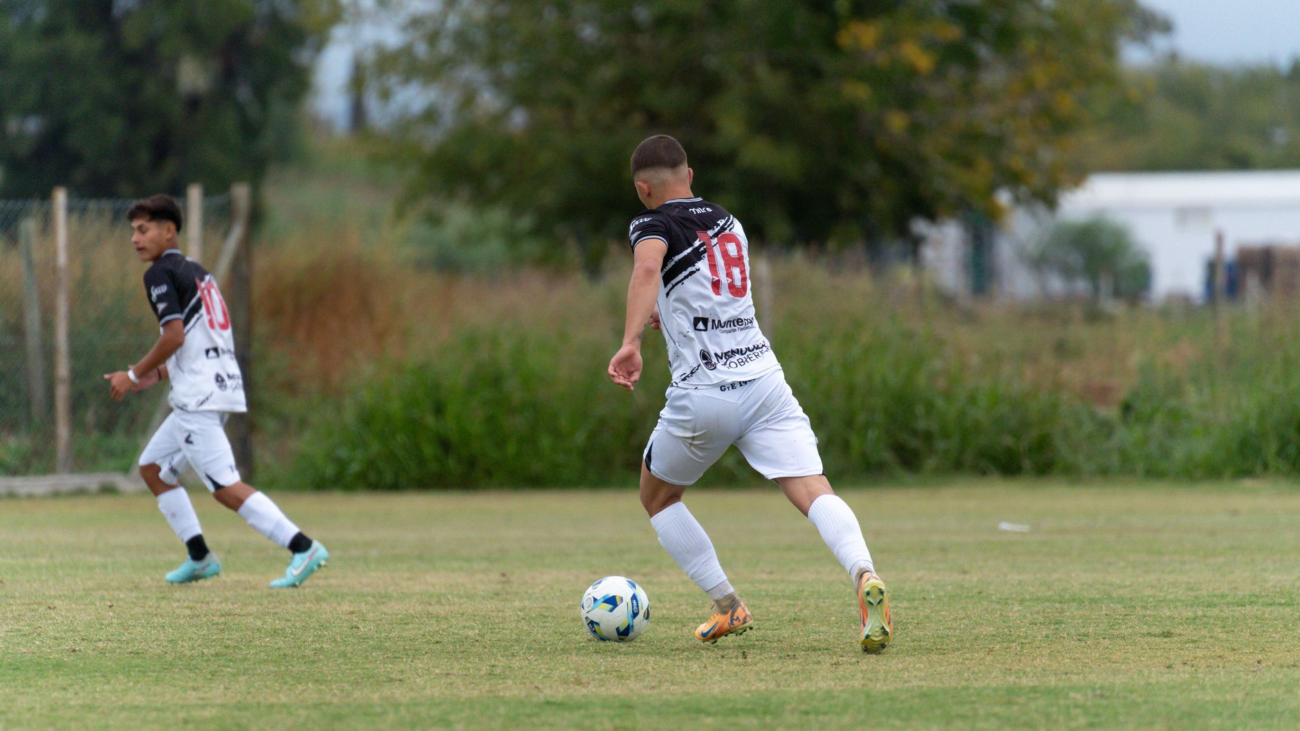 Two soccer players in action on a grassy field. One dribbles the ball as the other follows on an outdoor setting.