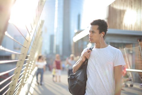 Free Serious young African American male in earphones and white shirt looking away and thinking while standing on street with large black bag on shoulder and listening to music against blurred urban environment in city Stock Photo