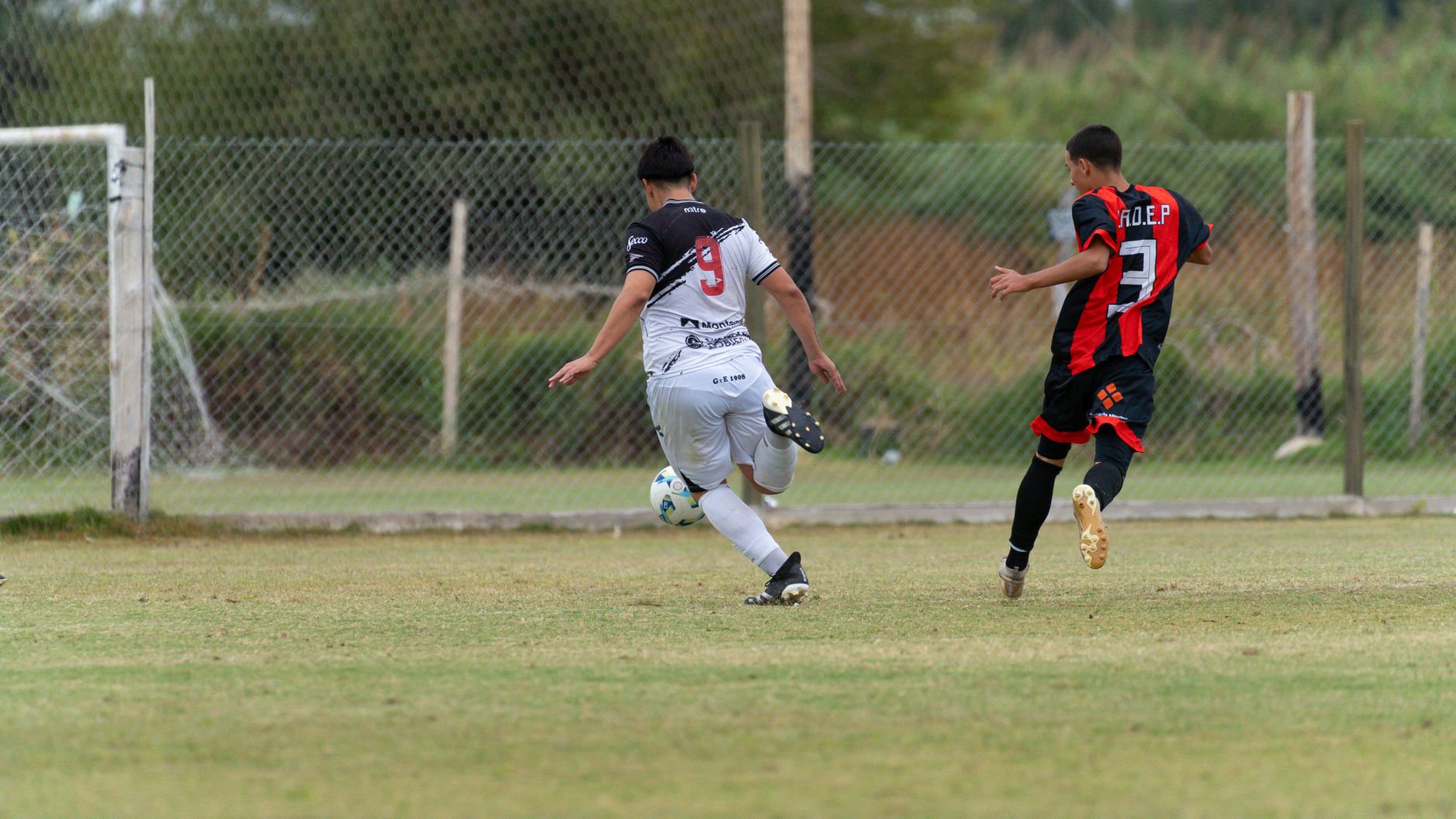 Two soccer players in action during a match on a grassy field with a fence background.