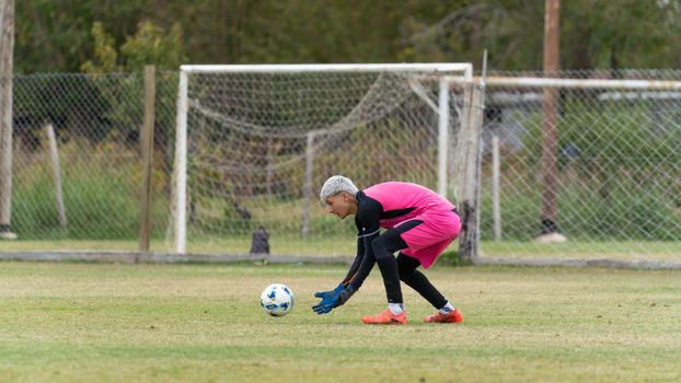 Goalkeeper in bright pink attire bending to catch a soccer ball during a game on a grassy field.