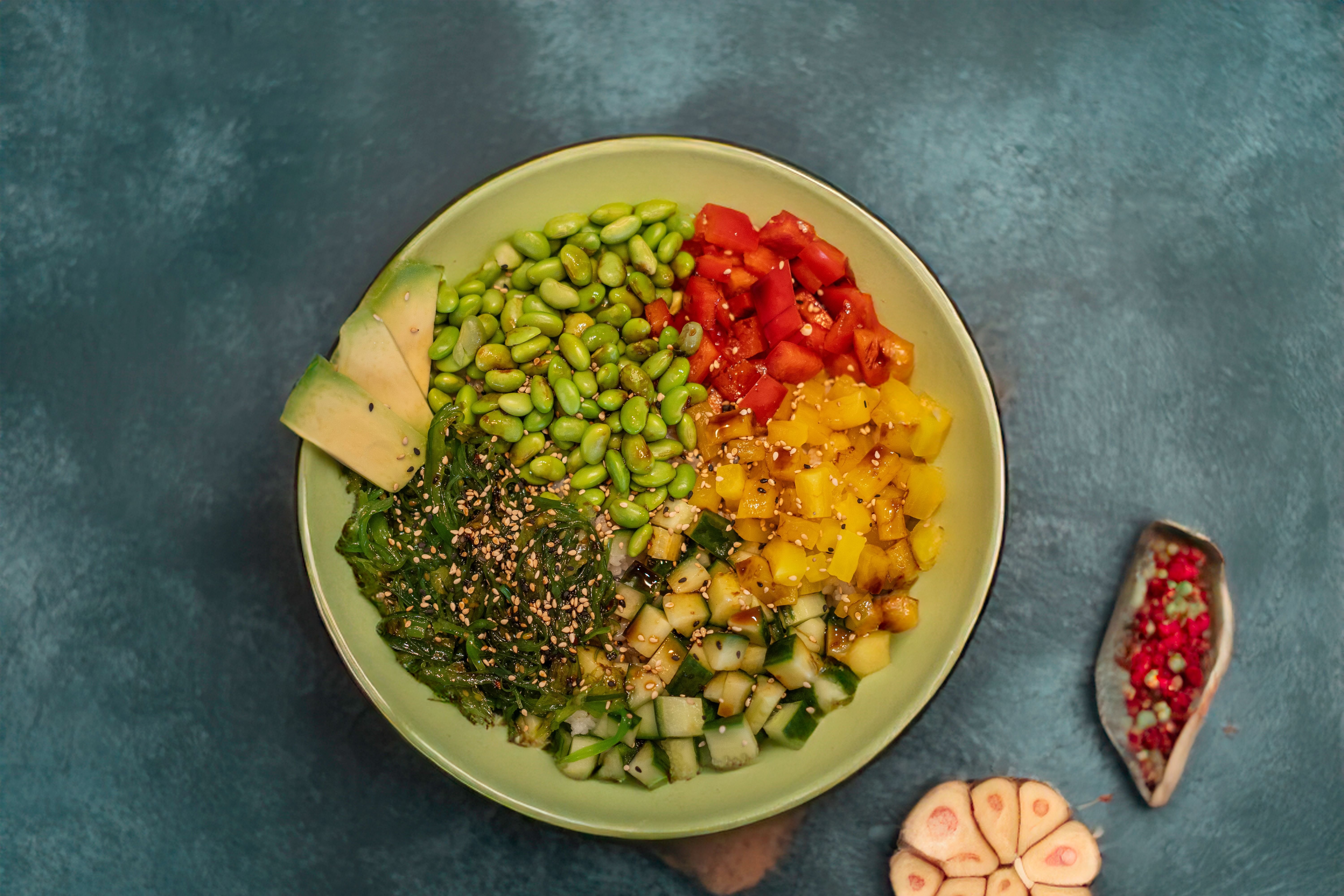 A colorful vegan poke bowl with fresh veggies, avocado, and spices on a textured blue background.