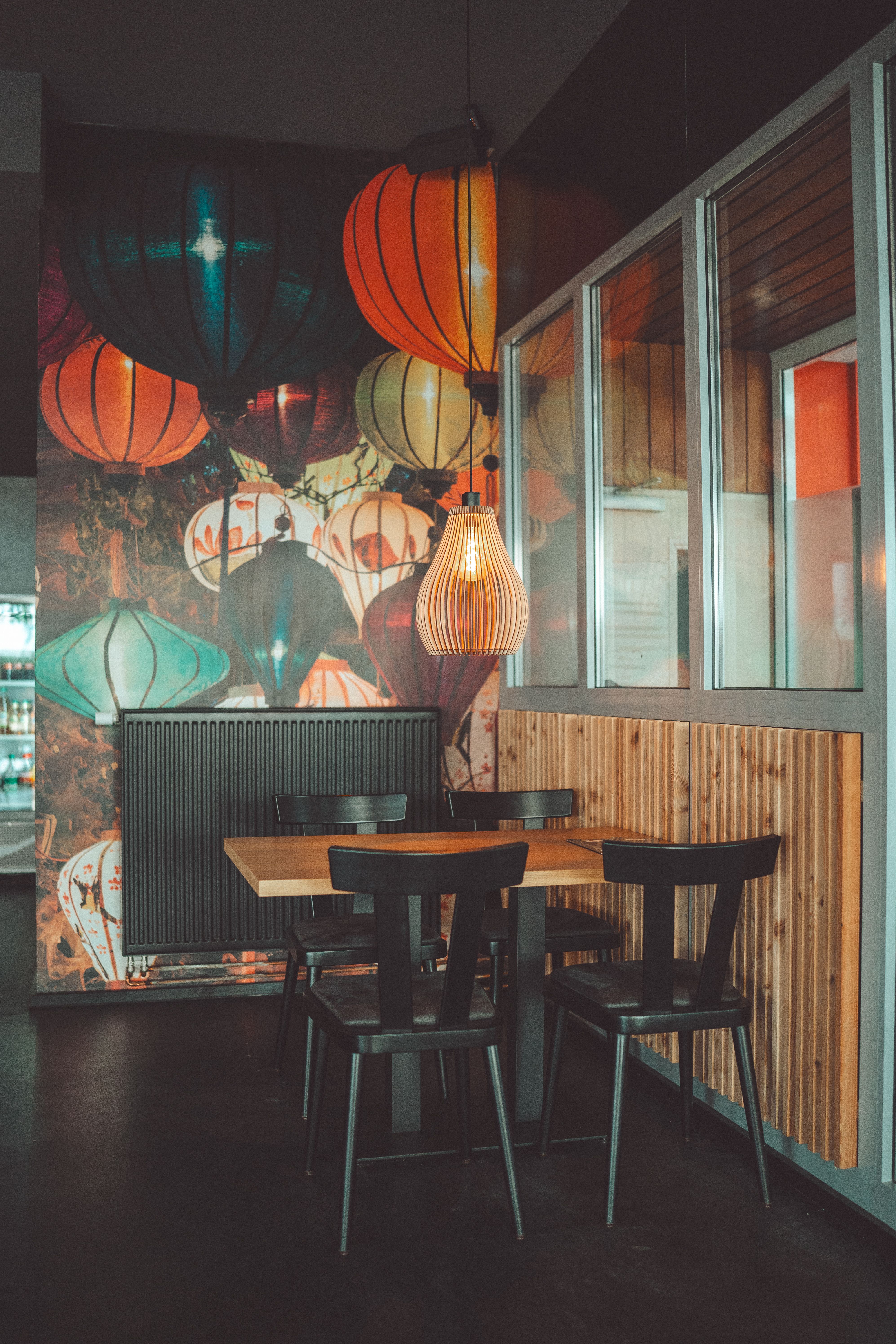 Coin de salle : table et chaises en bois sombre sous une suspension, devant une grande fresque de lanternes colorées